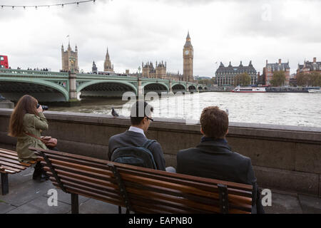 Zwei Männer in Anzügen und eine weibliche Touristen sitzen auf Bänken auf an der Themse, gegenüber des Palace of Westminster Stockfoto