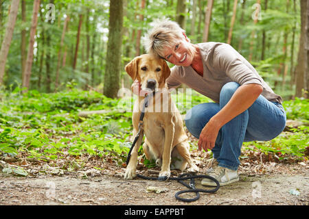 Glückliche ältere Frau sitzt mit Labrador Retriever in einem Wald Stockfoto