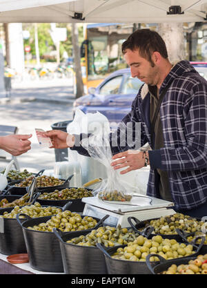 Grüne Oliven verkauft. Eine Olivenöle Anbieter in einem Straßenmarkt nimmt eine 10-Euro-Schein für einen Beutel mit grünen eingelegten Oliven, die auf einer Skala Stockfoto