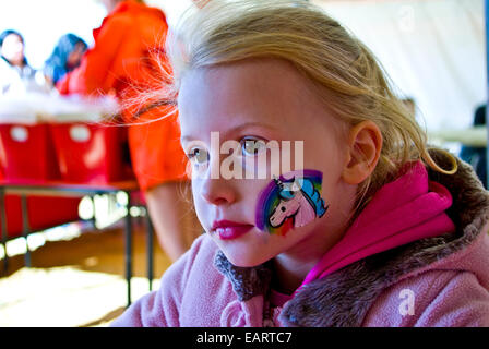 Ein blondes Mädchen mit einem Einhorn und Regenbogen gemalt auf die Wange. Stockfoto