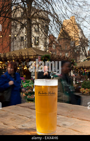 Ein erfrischendes Bier in ein graviertes Glas mit Logo "Manchester Markets" und Shopper im Hintergrund Manchester, UK Stockfoto