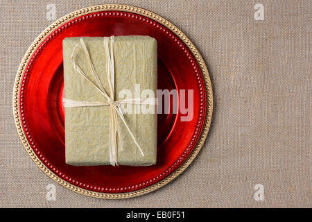 Ein gold Seidenpapier eingewickelt Weihnachtsgeschenk auf einem roten Ladegerät. Die Gegenwart wird mit Bast gebunden und beide Elemente sind auf einem Sackleinen-s Stockfoto