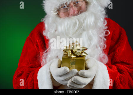 Nahaufnahme von Santa Claus ein kleines Weihnachtsgeschenk verpacktes in den Händen hält. Querformat auf Licht, dunkelgrünen Hintergrund Stockfoto