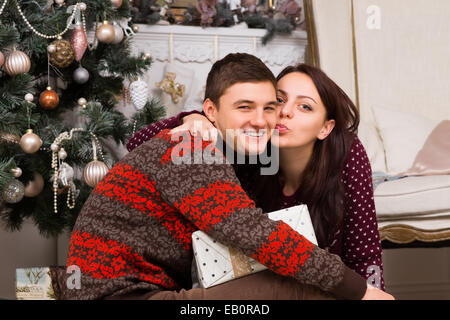 Junge Frau küsst ihren Freund um ihm für seine Weihnachts-Geschenk zu danken, wie sie auf dem Boden vor dem Weihnachtsbaum sitzen Stockfoto