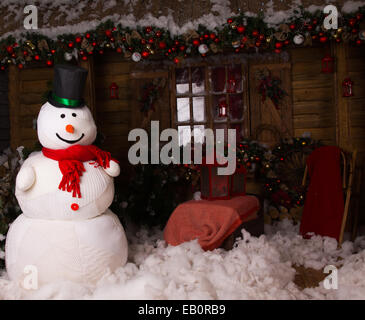 Großes Winter Schneemann mit Mütze am eingerichteten Holzhaus mit Baumwolle Schnee auf dem Boden stehen. Stockfoto