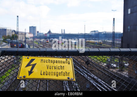 Gelbes Schild mit Hochspannung mit dem Bahnhof im Hintergrund Stockfoto