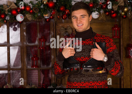 Fröhliche junger Mann trägt einen Pullover mit saisonalen Muster halten ein paar Schlittschuhen vor einem Chalet, geschmückt mit Christbaumkugeln und Girlanden, Konzept der Winterferien. Stockfoto