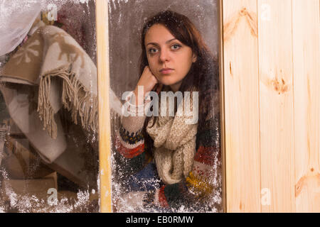 Traurige junge Frau in ein Winter-Kabine zu Weihnachten starrte mürrisch durch eine mattierte Holzfenster mit einem Blick von Einsamkeit und Sehnsucht. Stockfoto