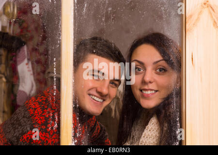 Schließen Sie glückliches junges Paar im Winter T-Shirts lächelnd hinter Glas Fenster während in die Kamera schaut. Stockfoto