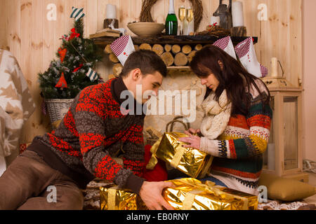 Junger Mann und Frau sitzen auf dem Boden in einer rustikalen Holzhütte eleganten metallic gold Weihnachtsgeschenke, die eifrig Lieferbindungen öffnen Stockfoto