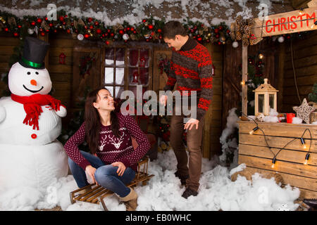 Paar tragen Pullover mit Schlitten draußen im Winter vor Blockhütte Stockfoto