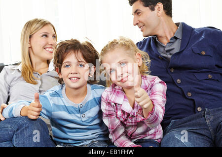 Zwei glückliche Kinder, die Daumen hochhalten, zwischen den Eltern auf dem sofa Stockfoto