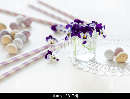 kleinen Blumenstrauß von Stiefmütterchen im Glas jar auf ein Deckchen auf weißen Tisch mit bunten Mini-Eiern und Pastell Strohhalme in Sonnenlicht getaucht Stockfoto