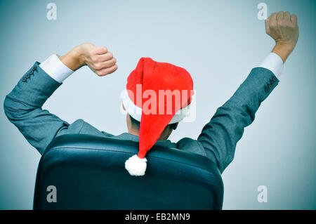 ein Mann in Anzug mit einer Nikolausmütze, streckte die Arme in seinem Bürostuhl Stockfoto