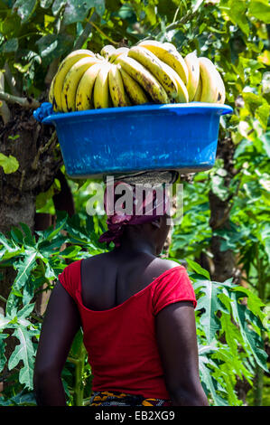 Eine Frau geht durch ein Dorf mit ein paar Bananen auf dem Kopf in einem Plastikeimer. Stockfoto