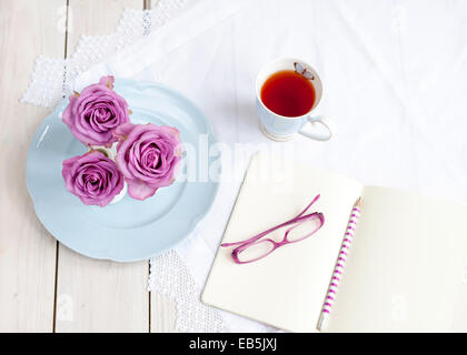 Drei Rosa Rosen in Vase, Becher Tee, blau Vintage Teller, Gläser, Notizbuch und Bleistift. Genießen Sie eine Tee-Pause Stockfoto