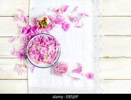 Schneiden Sie blass rosa Pfingstrosen und Blütenblätter in Schüssel weiß auf weiß Vintage spitzen Tischdecke auf Holztisch Stockfoto