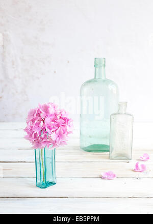 drei Vintage Glasflaschen mit Rosa Hortensie Blume Wand strukturiertes Pflaster auf weißer Holztisch Stockfoto