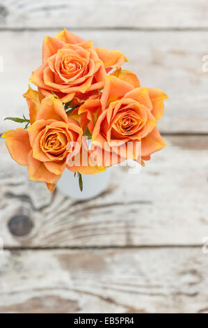 Drei orange Rosen in einer weißen Flasche auf einem notleidenden Holztisch Stockfoto