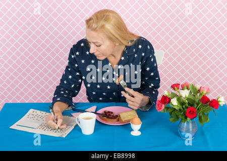 Reife Frau macht Kreuzworträtsel in Zeitungen beim Frühstück Stockfoto