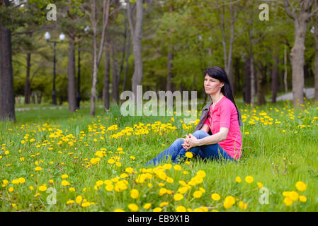 Junge Frau sitzt im Löwenzahn auf der Wiese und nachdenklich in die Ferne schauen Stockfoto