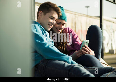 Teenager-Mädchen und jungen sitzen auf Boden, lächelnd und mit Blick auf Handy zusammen, Mannheim, Deutschland Stockfoto