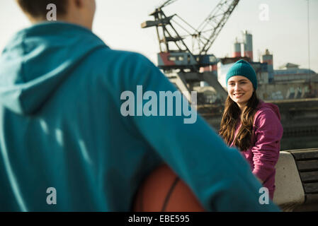 Teenager-Mädchen im Freien tragen Hauben, lächelnd und mit Blick auf Teenager halten Basketball, Industriegebiet, Mannheim, Deutschland Stockfoto