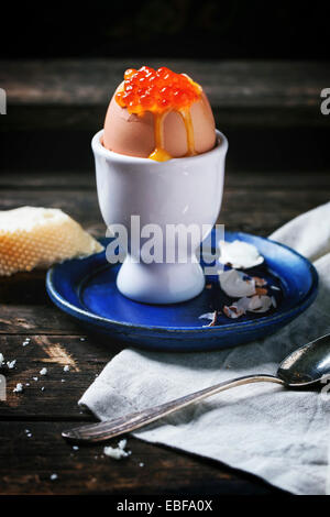 Frühstück mit wachsweiches Ei mit rotem Kaviar in weiß Eierbecher über alten Holztisch mit Brot serviert. Serien ansehen Stockfoto