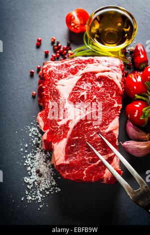 Rohes Rindfleischsteak mit Fleischgabel und Zutaten auf einem dunklen Hintergrund Schiefer Stockfoto