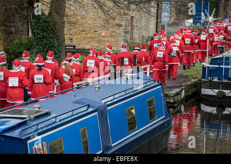 Ansicht der Rückseite des Menschen (Männer, Frauen und Kinder) in rot & weiß Vater Weihnachten Outfits gekleidet Gehen, Joggen, Laufen Vergangenheit Kanal Boote & die Teilnahme an der großen Skipton Santa Fun Run, eine jährliche fundraising Charity Rennen vom Rotary Club - Federn Zweig von Leeds Liverpool Canal Leinpfad, Skipton Stadtzentrum, dem North Yorkshire, England, Großbritannien organisiert. Stockfoto