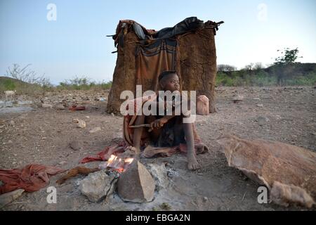 Junge Schäferin aus der Himba Leute sitzen am Lagerfeuer vor ihrer Hütte Ombombo, Kaokoland, Kunene, Namibia Stockfoto