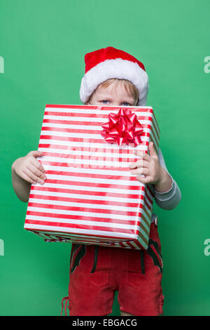 Kleines Kind im roten Kostüm Zwerg versteckt sich hinter roten Geschenkbox. Studio-Porträt auf grünem Hintergrund Stockfoto