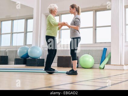 Weibliche Trainer helfen senior Frau auf ein Balance Board im Fitnessstudio. Ältere Frau, die Ausübung von Trainer unterstützt. Stockfoto