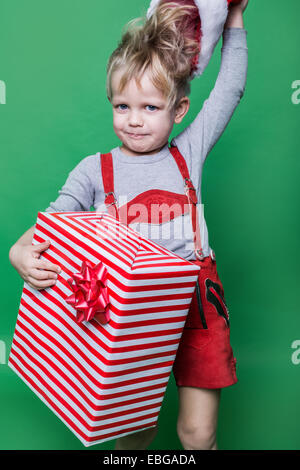 Kind in Rotes Kostüm Zwerg hält Weihnachten Geschenk und wirft Weihnachtsmann-Mütze. Ungezogenes Kind Stockfoto