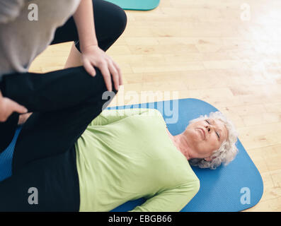 Ältere Frau liegt auf Gymnastikmatte mit ihrem persönlichen Trainer, ihr zu helfen tun Bein Strecken im Fitnessstudio. Ältere Frau dehnen. Stockfoto