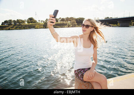 Junge Frau sitzt im Riverside Smartphone Selfie, Donauinsel, Wien, Österreich Stockfoto