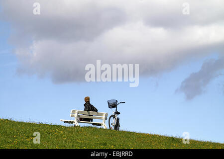 Frau sitzt auf einer Bank am Deich, Fahrrad neben ihr, Deutschland, Niedersachsen, Cuxhaven Stockfoto