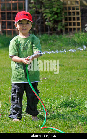 kleiner Junge stehen auf Wiese und plantschen mit Gartenschlauch Stockfoto