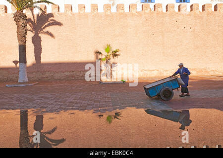 Mann mit einem Karren an der historischen Stadtmauer widerspiegelt in Pfütze Wasser nach einem Regenschauer, Marokko Essaouira Stockfoto