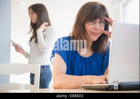 Mutter mit Laptop, Tochter (14-15) mit Handy im Hintergrund Stockfoto