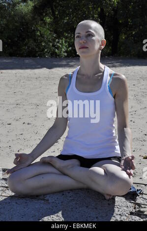 Glatze Frau im Lotussitz sitzen an einem Strand, Kopf gegen dunkle Bäume Stockfoto