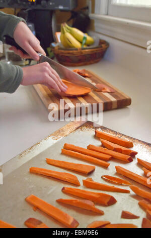 Junge Frau Hände geschnitten Süßkartoffel Pommes Frites auf ein Schneidebrett mit rohem Pommes auf ein Backblech im Vordergrund Stockfoto