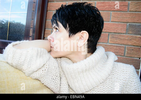 Schöne junge Frau saß in einem Sessel durch ein Fenster tief in Gedanken, Nachdenken, Träumen, den Blick in den Raum, der aus einem Fenster Stockfoto