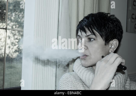 Schöne kurze, dunkelhaarige Frau vaping und durch ein Fenster tief in Gedanken Stockfoto