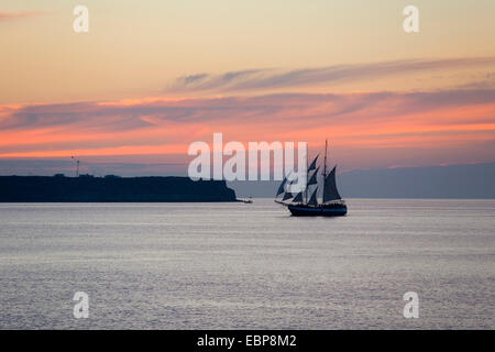 IA, Santorini, südliche Ägäis, Griechenland. Segelschiff, vorbei an der Insel Thirasia, Dämmerung, rosa Wolken im Himmel. Stockfoto