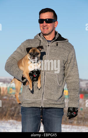 Ein Mann hält seinen Bugg-Hund (Kreuzung Boston Terrier und Pug) im Stadtpark mit klarem blauem Himmel Stockfoto