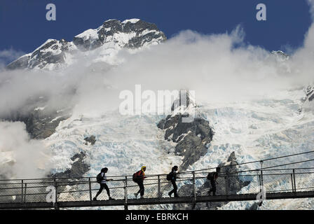 Touristen, die Überquerung einer Hängebrücke über den Fluss Hooker im Hooker Valley mit den vergletscherten Mt. Sefton in den Hintergrund, Neuseeland, Südinsel, Mount Cook Nationalpark Stockfoto