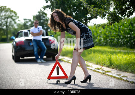 junge Frau Aufstellen eines Warndreiecks mit einem Lächeln nach einer Panne auf einer Landstraße während der Einnahme es humorvoll, Deutschland Stockfoto