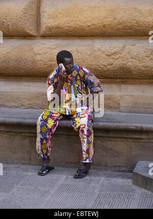 Ein Afro-Karibischen Mann in einem bunt gemusterten Outfit sitzt auf einer Steinbank in Florenz sprechen auf dem Handy Stockfoto