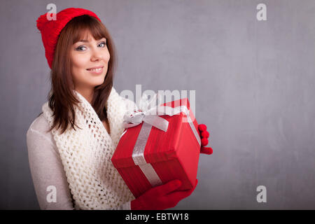 Junge Frau in roten Mütze und Handschuhe hält Weihnachtsgeschenk in rotem Papier gewickelt, Stockfoto